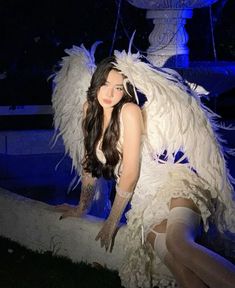 a woman dressed in white posing with an angel wings on her head and legs, next to a fountain at night