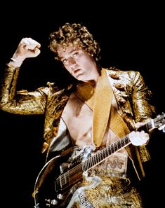 a man in gold jacket holding a guitar and pointing to the side with his right hand