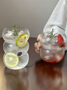 two glasses filled with ice and fruit on top of a wooden table next to each other