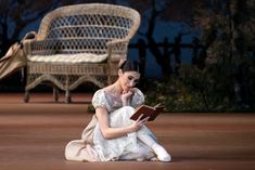 a woman sitting on the ground reading a book in front of a wicker chair