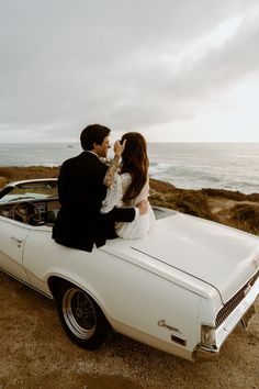 a bride and groom sitting on the hood of a white car in front of the ocean