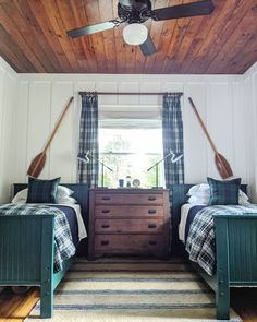 two beds in a room with wooden ceilings and plaid blankets on the bedding, under a ceiling fan