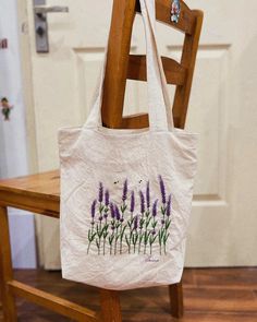 a white tote bag with purple flowers painted on it sitting on a wooden chair