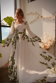 a woman in a long white dress with green leaves on it standing next to a window