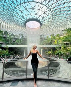 a woman in a black dress is standing on a balcony near a fountain and trees