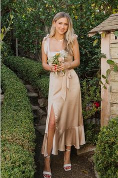 a woman in a dress holding flowers and standing next to a garden area with bushes