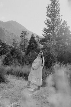 black and white photograph of a pregnant woman standing in front of some trees with mountains in the background