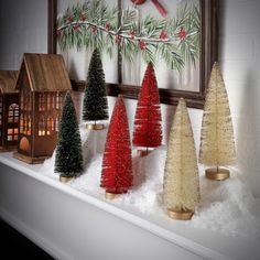 small christmas trees are lined up on a mantel in front of a framed holiday scene