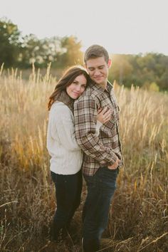 a man and woman standing in tall grass with their arms around each other as the sun sets behind them