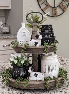 three tiered tray with jars and plants in them on a kitchen counter next to a clock