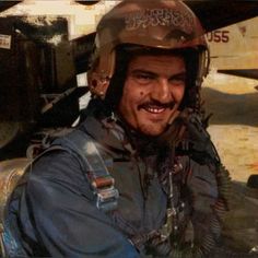 a man with a helmet on sitting in front of an old fighter plane and smiling at the camera