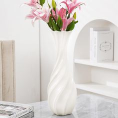 a white vase filled with pink flowers on top of a table next to a book