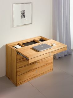 a wooden desk with a laptop on it in front of a white wall and window