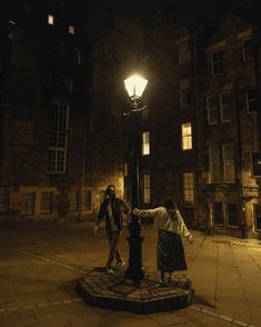 two people standing on the sidewalk near a lamp post in an old city at night