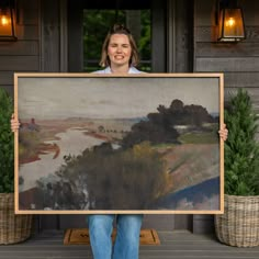 a woman holding up a large painting in front of a house with potted plants