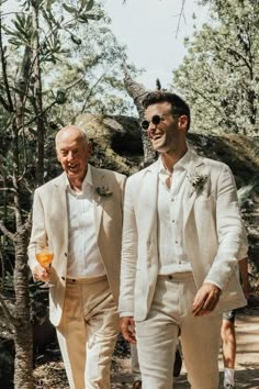 two men in white suits walking down a path with one holding a drink and the other wearing sunglasses