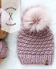 a knitted hat with a pom - pom sits next to balls of yarn