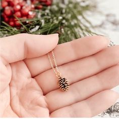 a hand holding a tiny pine cone necklace in front of a christmas tree with berries