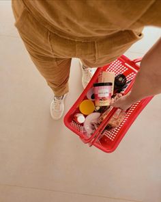 a person standing next to a red shopping basket filled with food and condiments