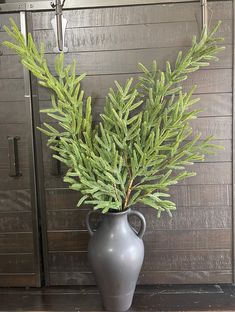 a gray vase with green branches in it sitting on a wooden floor next to a wall