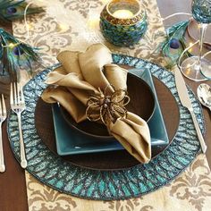 a place setting with peacock napkins and silverware on a wooden tablecloth covered dining room table