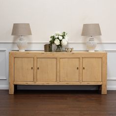 two lamps on top of a wooden sideboard with flowers in vases next to it