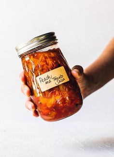 a hand holding a jar filled with pickle and tomato sauce on a white surface