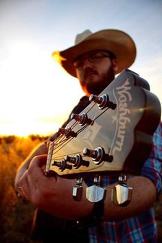 a man wearing a cowboy hat and holding an acoustic guitar in his hands with the sun setting behind him