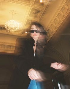 a woman in black jacket and sunglasses sitting on a chair next to a chandelier