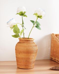 three white roses in a woven vase on a wooden table next to a wicker basket