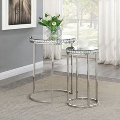 two glass tables with metal legs in front of a gray wall and white drapes