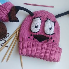 a pink knitted hat with eyes and nose sitting next to knitting needles, balls of yarn and crochet hooks