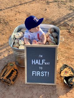 a little boy sitting in a bucket filled with baseballs and mitts next to a sign that says i'm half way to first