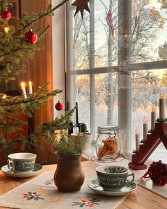 a christmas tree in front of a window with tea cups and saucers on the table