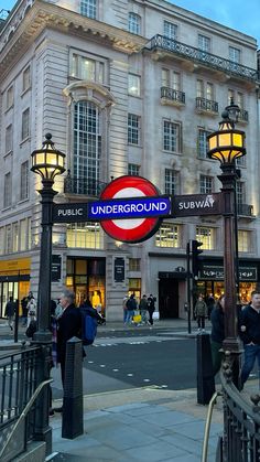 people are standing on the sidewalk in front of an underground subway sign and lampposts