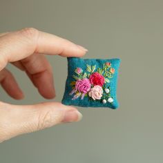 a hand is holding a small embroidered flowered square pillow with flowers on it's side