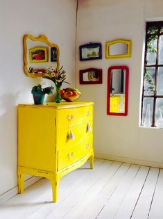 a yellow dresser in a white room with pictures on the wall and flowers sitting on top