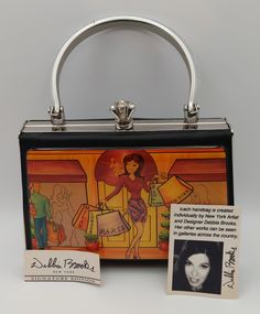 purse is a Debbie Brooks Signature Edition piece, created individually by the New York artist and designer. The front panel features a vibrant, stylized cityscape with figures engaged in shopping, highlighting a central figure holding bags labeled "PARIS." The background blends warm colors, giving a bustling city vibe. Debbie Brooks' works are showcased in galleries across the country, making this purse part of a limited-edition collection, which could be appealing to collectors and admirers of her art. Debbie Brooks' Signature Edition purses, like the one you have, were often released in the early 2000s. For example, some of her limited-edition pieces were created for events like the 2004 Tony Awards. Given the style and design, it's likely that your purse was made around that time period Artistic Rectangular Evening Bag, Bustling City, City Vibe, Tony Awards, Evening Clutch, Chic Boutique, Handbag Purse, New Yorker, Clutch Handbag