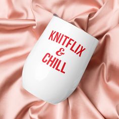 a white coffee mug with the words knitflix and chill printed on it sitting on a pink satin surface