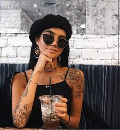 a woman sitting at a table with a drink in her hand and tattoos on her arm