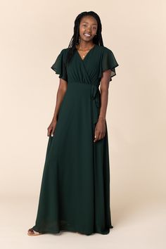 a woman in a dark green dress smiles at the camera while standing against a beige background