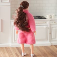 a doll in a pink outfit standing on a wooden floor next to a white kitchen