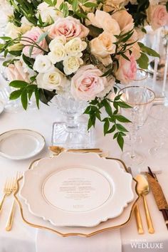 the table is set with white and gold plates, silverware, and pink flowers