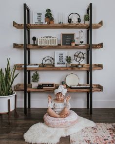 a baby sitting on a rug in front of a book shelf with books and plants