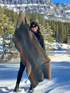 a woman standing in the snow holding up a green and brown knitted shawl