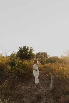 a woman in a white dress is standing on a dirt path near bushes and trees