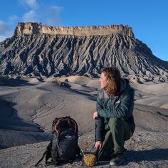 a woman sitting on top of a mountain next to a backpack and food container in front of a large rock formation