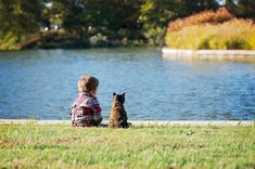 a little boy sitting next to a dog on the grass near a body of water