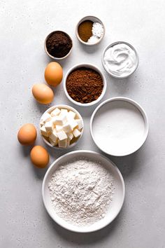 an assortment of ingredients are shown in bowls on a white counter top, including eggs and flour