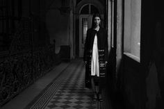 black and white photo of woman in long coat standing by door with checkered floor